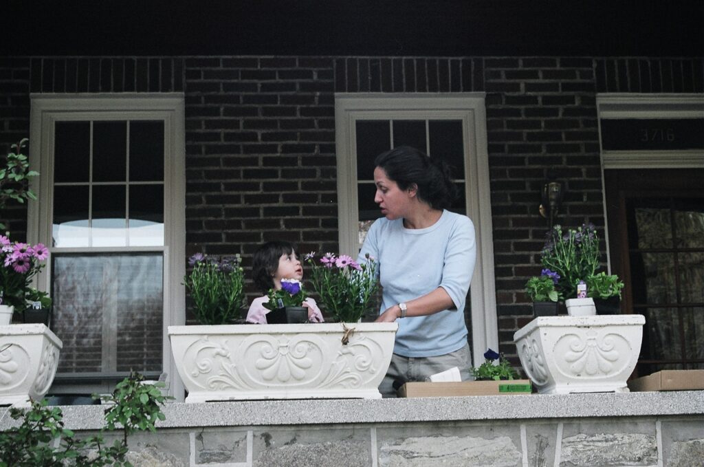 A mother and her daughter plant flowers in a planter on their front porch.