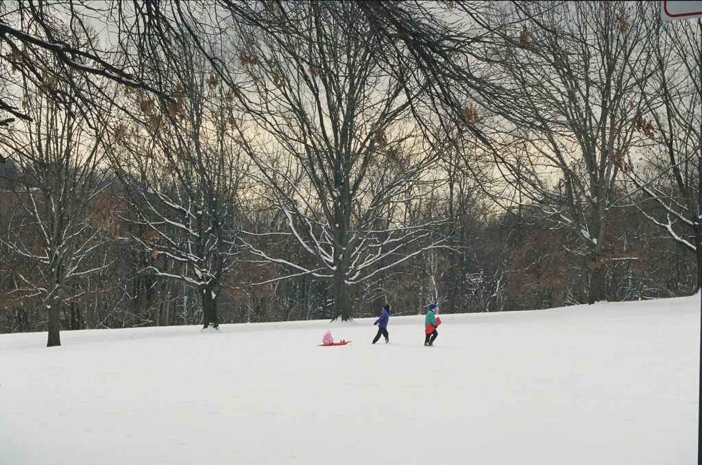 Park covered in snow. Three figures, one pulled in a sled.