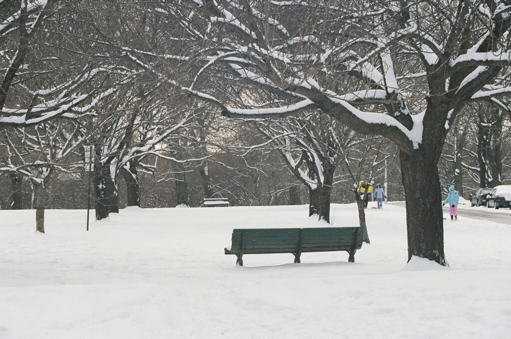 Bench in the park covered in snow. Sledders walking by.