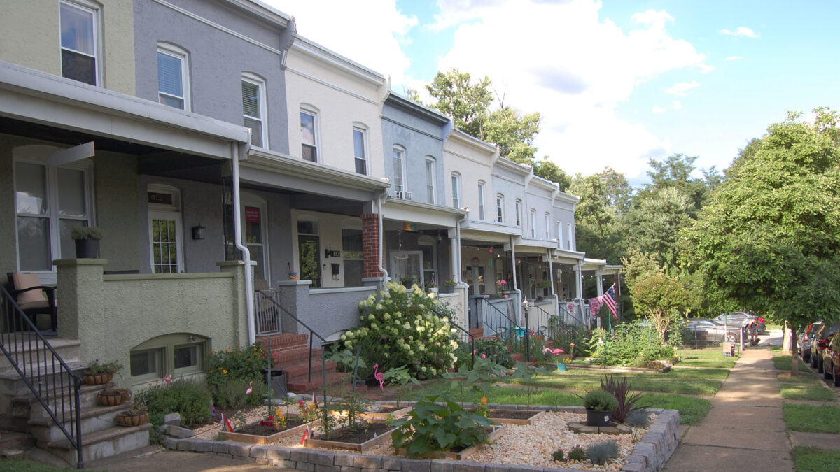 A row of painted stucco rowhouses with gardens in front, and a sidewalk. The park can be seen in the background.