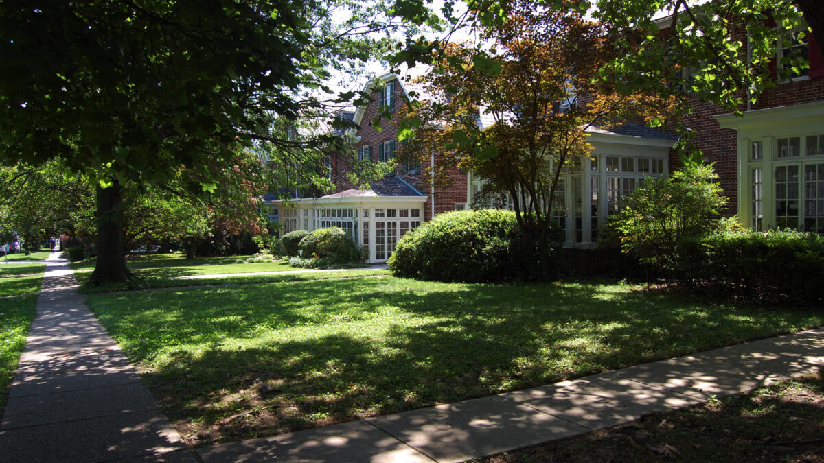 Semi detached colonial style brick row homes with sun rooms. Green yards with trees in front.