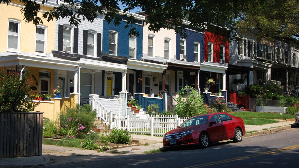 Multi-colored stucco rowhouses with landscaped gardens in front. The street in front is lined with trees.