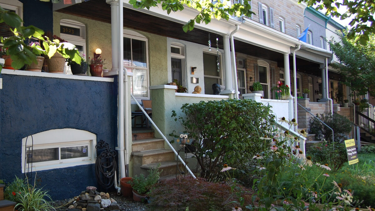 A line of variously-colored stucco and formstone rowhouses with steps leading up to large front porches. Gardens in front are in summer bloom.