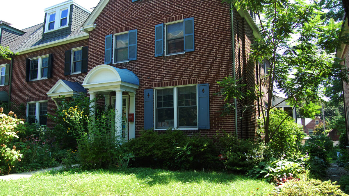 A wide brick corner house of a row grouping. Small porch with rounded portico. Green lawn in front with flowery and green landscaping.