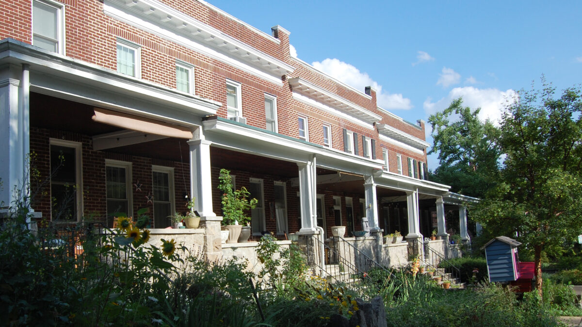 Line of brick rowhouses with stone front porches and blooming gardens in front. One house has two little free library houses in the yard--one for adults and one for children.