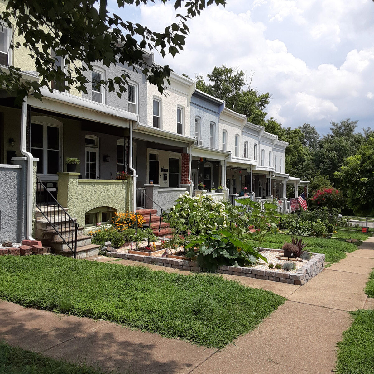 Multi-colored stucco rowhomes with covered porches. Many flowers bloom in the front gardens. Wyman Park can be seen in the background.
