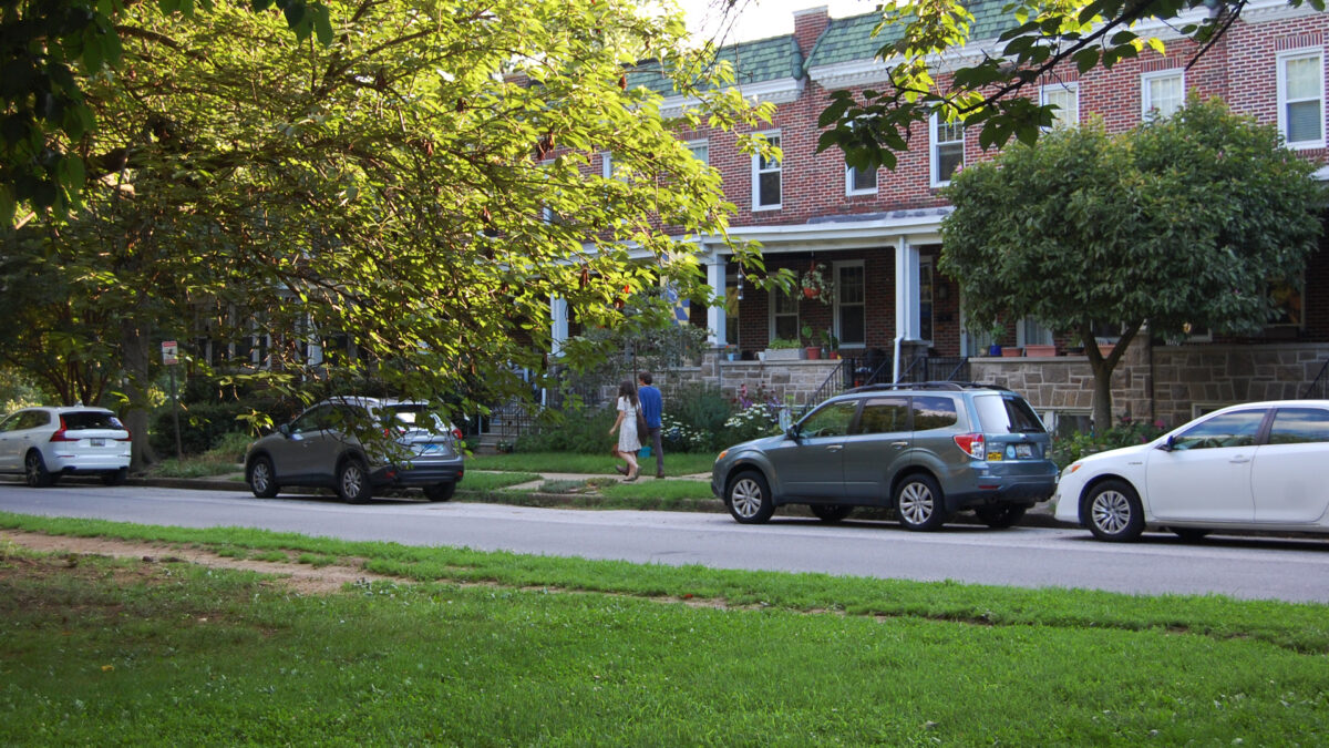 Two pedestrians enjoying a walk on a sidewalk across from the park on a summer day.